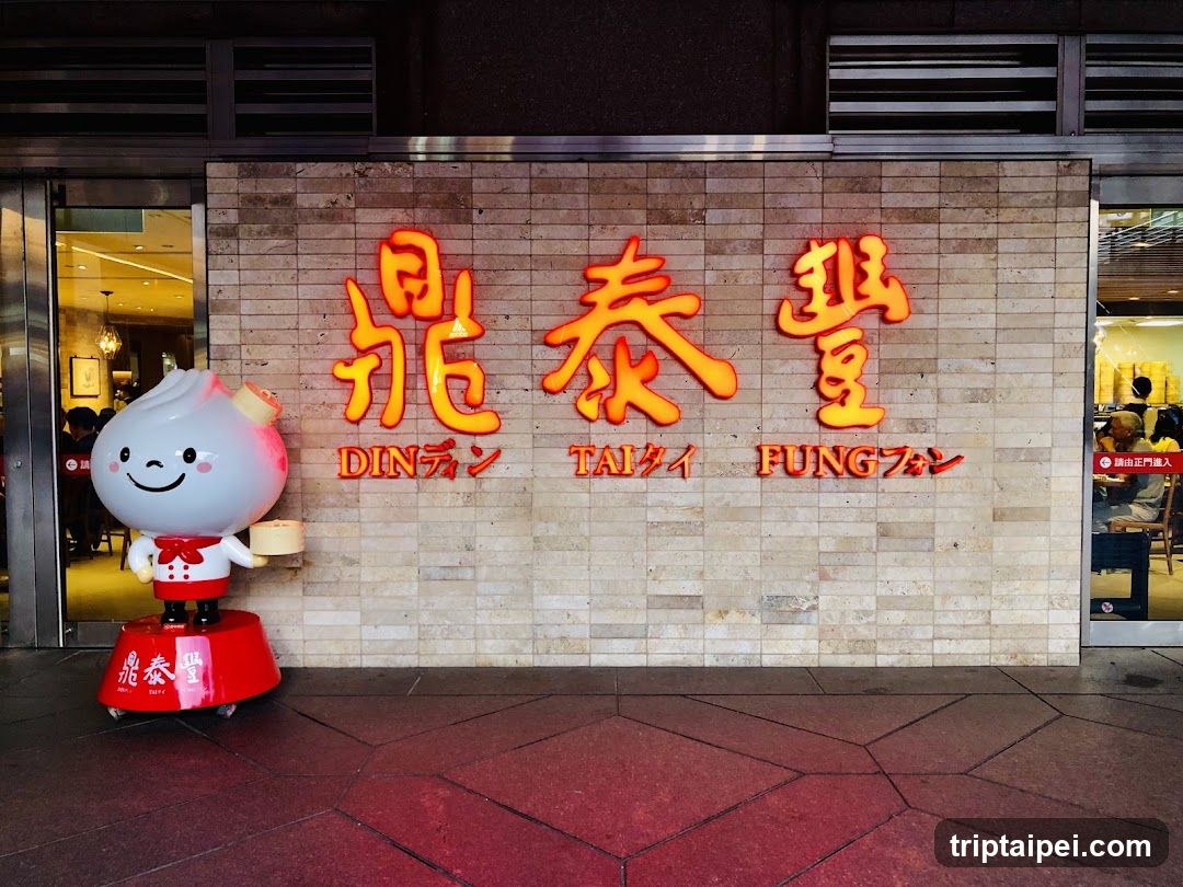 Photo of the exterior entrance showing a tiled wall with illuminated Chinese characters and English transliteration 'DIN TAI FUNG'. A cartoon dumpling mascot stands on a red base to the left. Glass doors reveal interior seating.