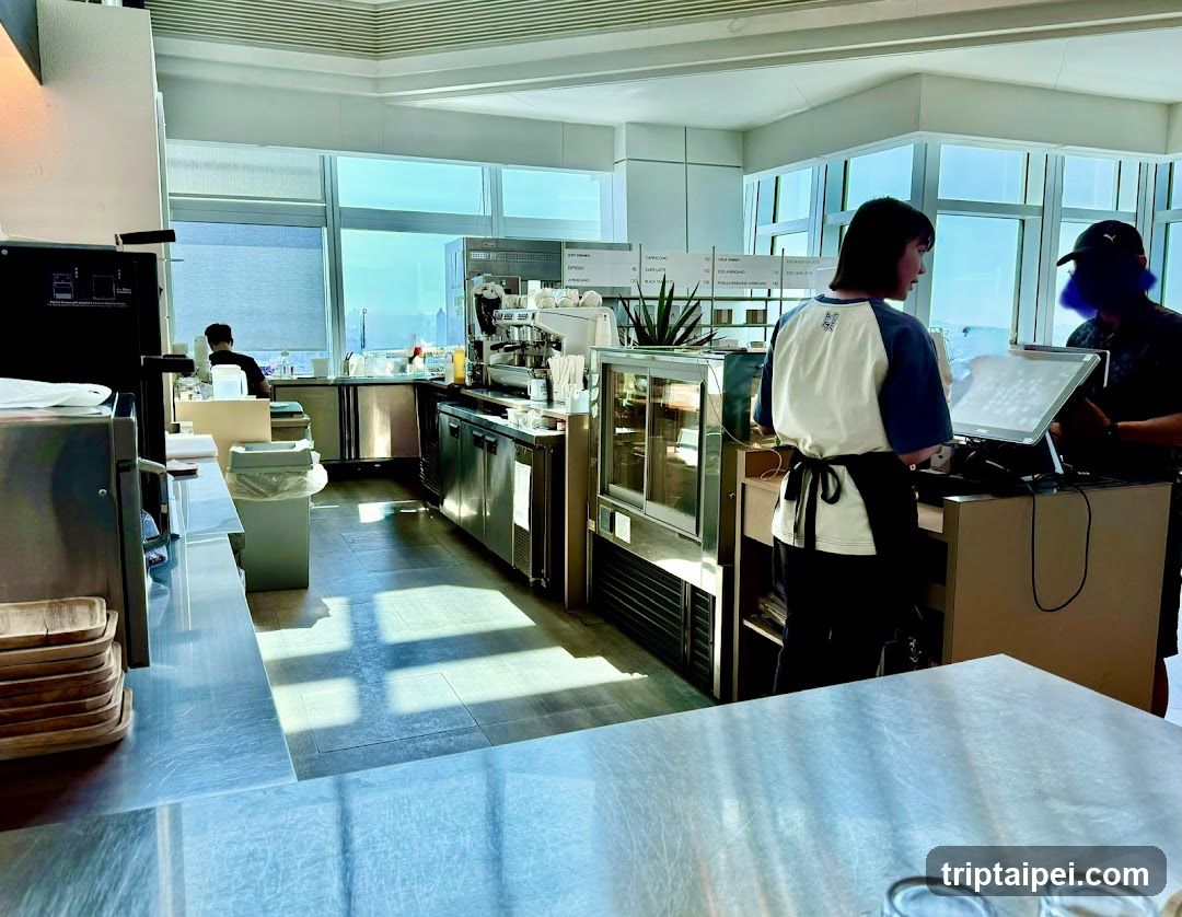 Interior view of a modern coffee shop featuring stainless steel equipment, large windows with natural light, and staff members working behind the counter. Visible items include espresso machines, display cases, and trays stacked on shelves.