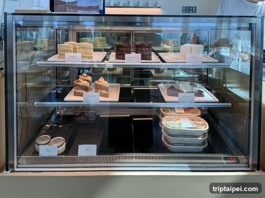A glass display case in a cafe or bakery, showcasing various pastries and cakes on multiple shelves. The top shelf features layered cakes and brownies with descriptive cards. Below are slices of cake, possibly cheesecake or sponge cake, and rectangular desserts. The bottom section contains packaged food items like sandwiches or wraps, along with small jars that might be jams or spreads. A digital temperature indicator is visible at the bottom right.
