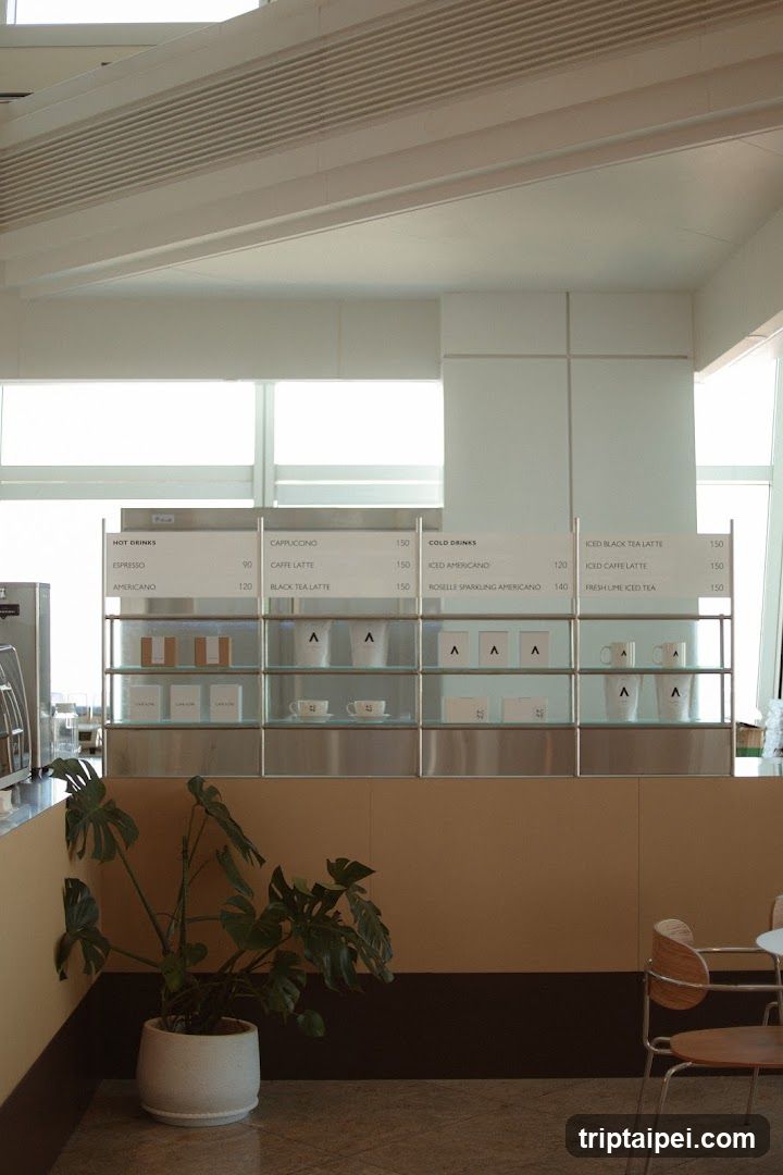 Interior view of a modern coffee shop featuring a glass display cabinet with menu boards and product samples. A potted plant sits in the foreground, and part of a seating area is visible on the right.