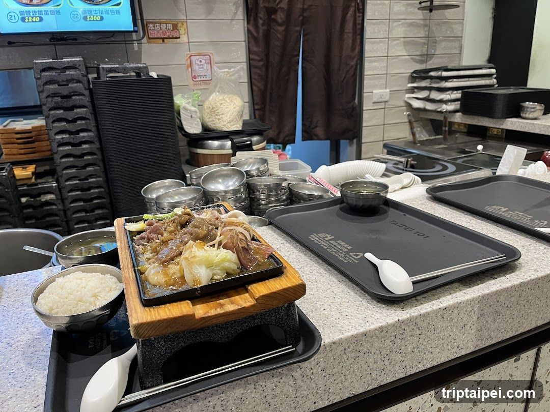 A food preparation or serving area in a restaurant, featuring a wooden tray with grilled meat and vegetables on top of rice, alongside stacked bowls, trays, and kitchen equipment.