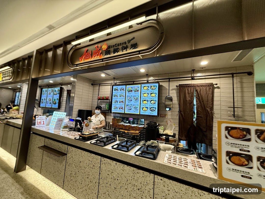 A food stall in a mall or shopping center, featuring a counter with cooking equipment, a staff member wearing a mask preparing food, multiple illuminated menu boards displaying dishes, promotional signs on the counter, and a brown curtain covering an entrance to the back area.