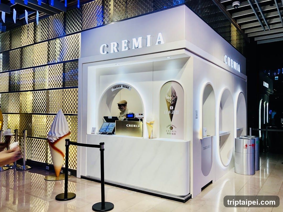 A modern, white ice cream shop with 'CREMIA' signage prominently displayed on the facade. The storefront features arched windows and a counter where staff are visible behind the glass. A large decorative ice cream cone stands outside near black stanchions. The background includes a golden textured wall.