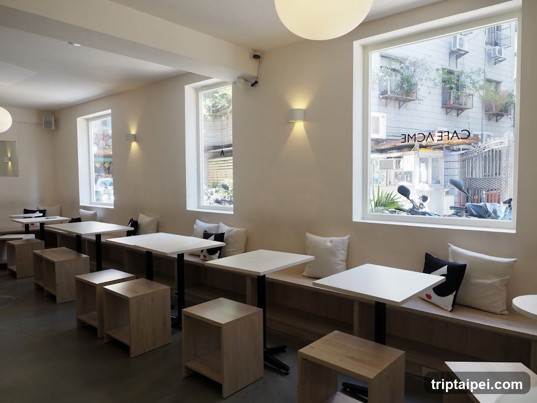 A modern cafe interior featuring light-colored wooden tables and stools, white cushions on built-in benches along the walls, and large windows offering a view of the street outside. The space is well-lit with wall-mounted lights and natural light from the windows.