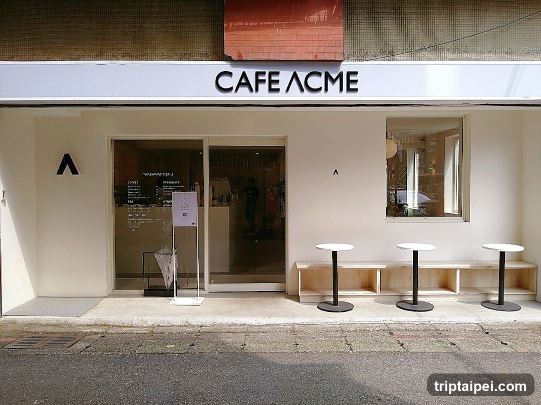 A modern cafe storefront with a clean white facade and black signage reading 'CAFE ACME'. The entrance features glass doors with menu boards inside, displaying coffee options like espresso and specialty drinks. Outside, there are three small round tables on black stands near a wooden bench. A minimalist logo is visible on the wall beside the door.
