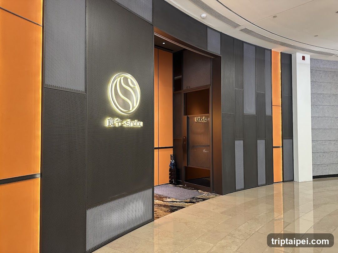 A modern storefront entrance featuring a dark perforated metal panel with an illuminated circular logo and Chinese characters below it. The building has orange vertical panels on the left, gray textured walls on the right, and a glass door leading inside. A patterned rug is visible at the threshold.