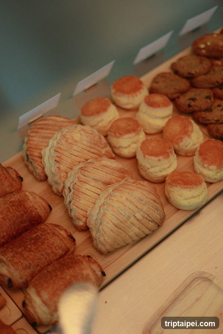 A close-up view of a wooden display case filled with various baked goods, including croissants, scones, and cookies. Small white labels are placed in front of each item.
