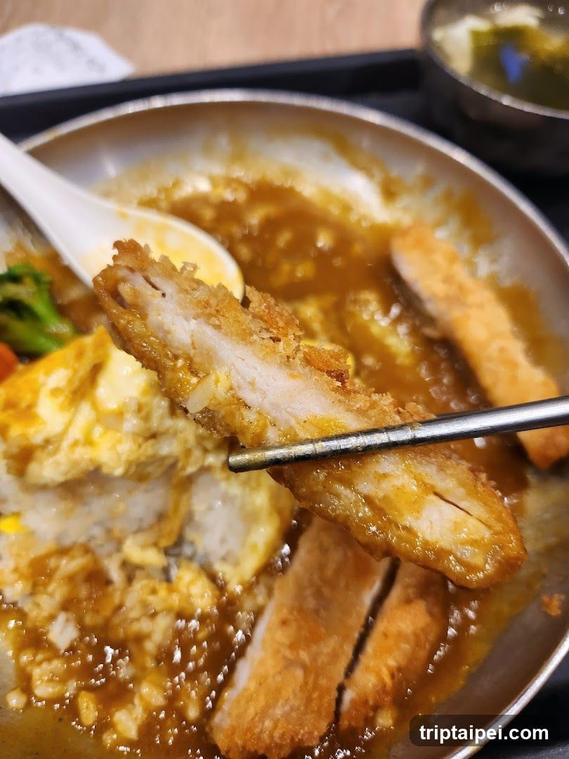A close-up of a bowl of curry rice featuring breaded cutlets, fried egg, and vegetables in rich brown sauce. Chopsticks hold up a piece of cutlet. A small side dish is visible in the background.
