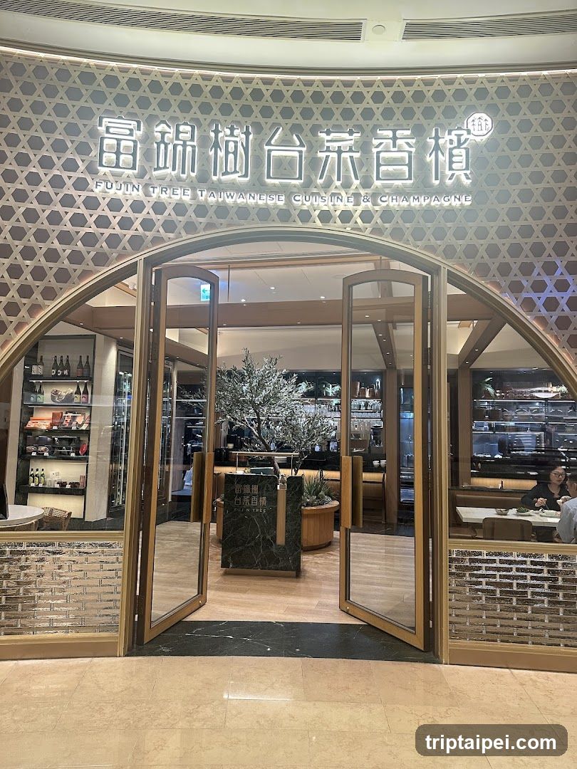 The image shows the storefront of a restaurant named 'Fujiin Tree Taiwanese Cuisine & Champagne'. The sign features illuminated Chinese characters and English text above an arched entrance with glass doors. Inside, there is a modern interior with wooden furniture, shelves displaying bottles, and a decorative tree centerpiece.
