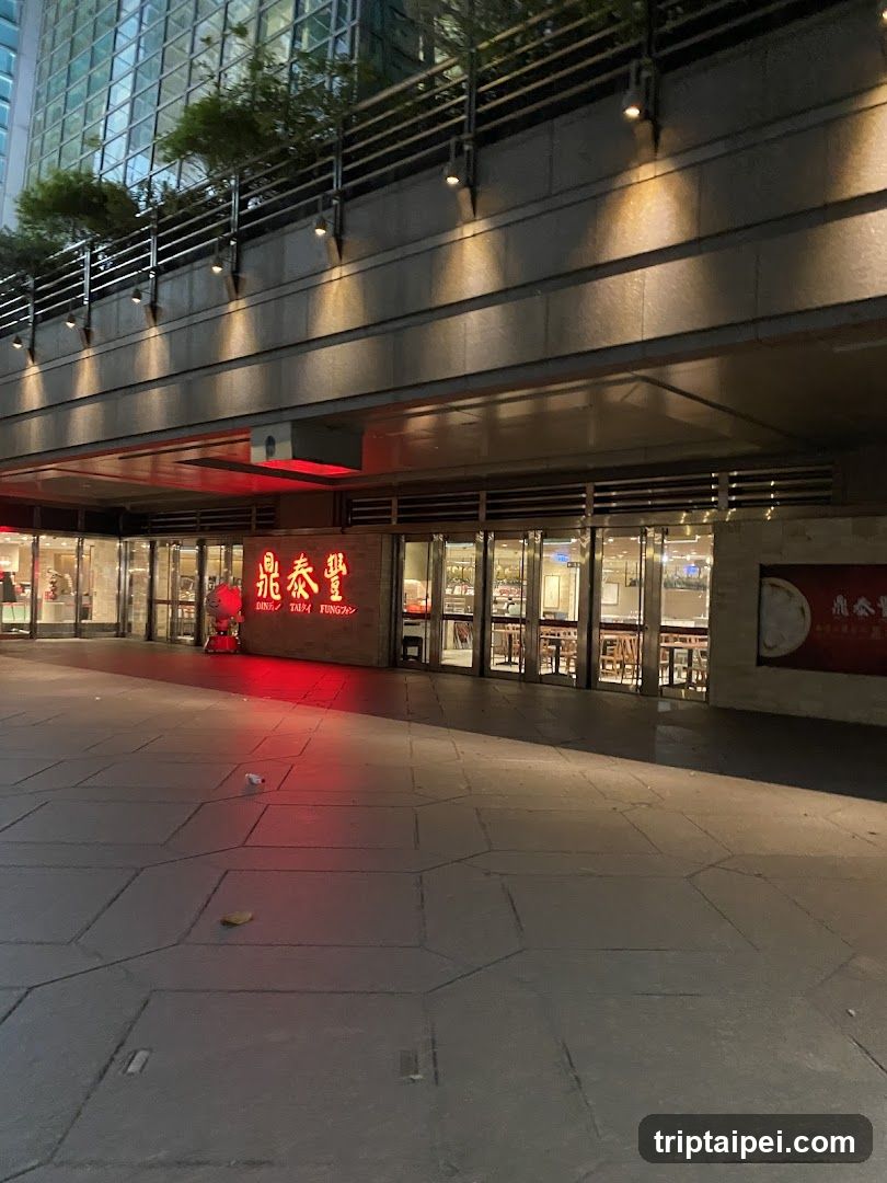 A nighttime view of a restaurant entrance with illuminated signage displaying Chinese characters and English text 'DINING'. The storefront features glass doors, warm interior lighting visible through windows, and modern architectural elements including metal railings above. A red light reflects on the pavement.
