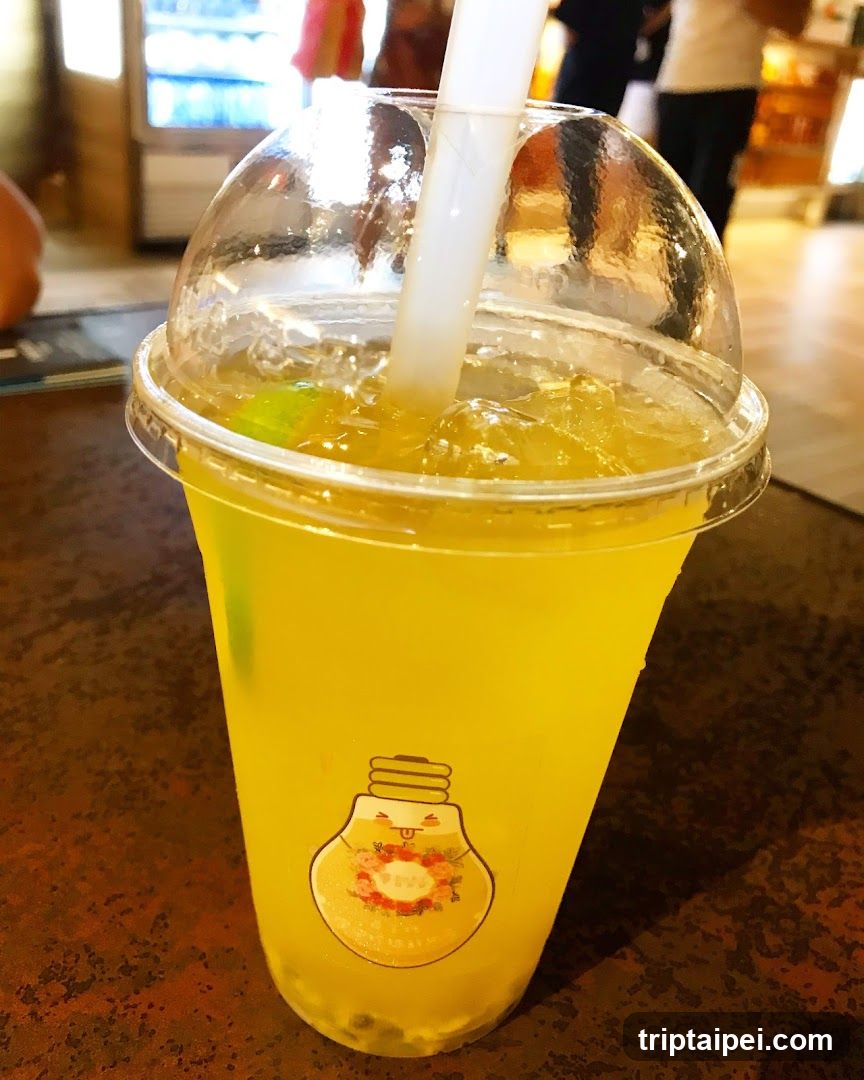 A close-up of a yellow iced beverage in a clear plastic cup with a dome lid and white straw. The cup features a lightbulb logo with floral design. Background shows blurred interior setting, possibly a cafe or shop.