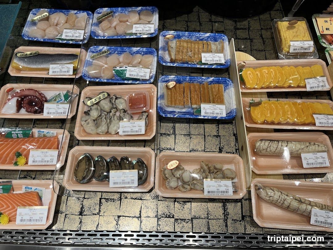 A refrigerated display case in a shop showcasing various fresh seafood items, including salmon sashimi, abalone, scallops, octopus, and other shellfish, arranged neatly on trays with price tags.