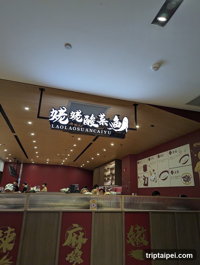 Interior shot of a restaurant with wooden ceiling and red walls. A sign reads '姥姥酸菜鱼 LAOLAO SUANCAIYU'. Staff are visible behind the counter, and menu boards display dishes on the wall.