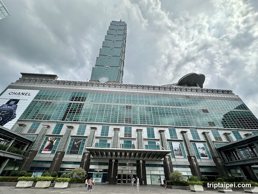 Low-angle shot of the Taipei 101 tower rising behind a large glass-fronted shopping center with visible Chanel signage, under cloudy skies.