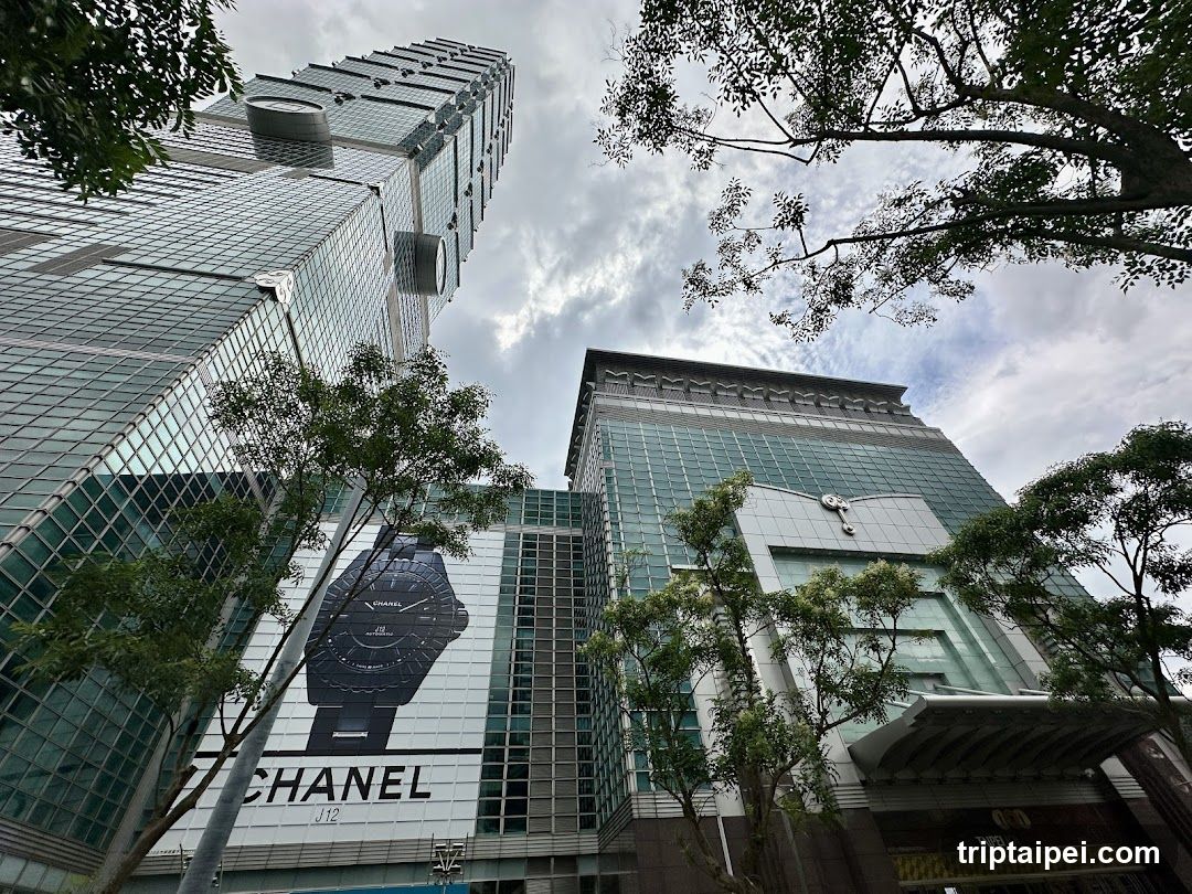 Low-angle shot of modern glass skyscrapers under cloudy sky. Large Chanel watch advertisement displayed on building exterior, with trees framing the scene.
