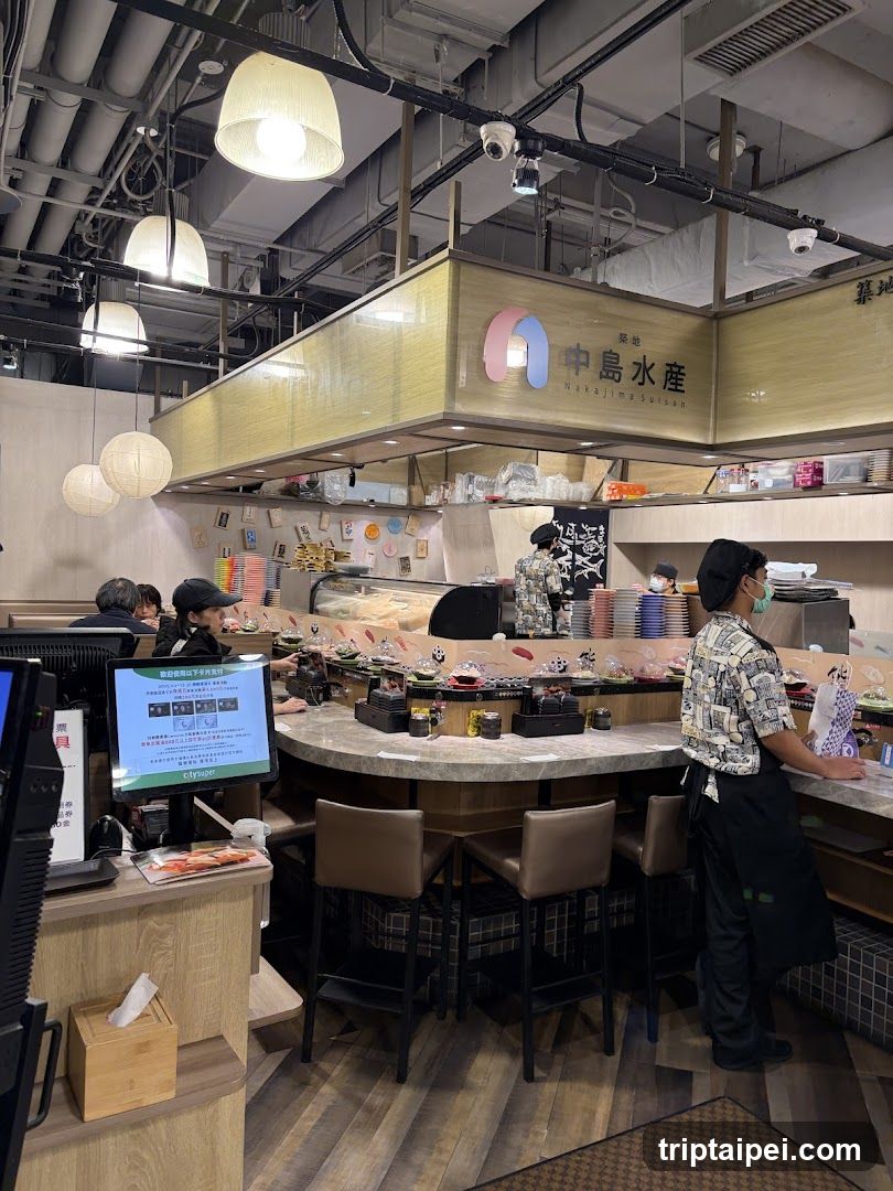 Interior view of a sushi restaurant with staff preparing food behind the counter, customers seated at tables, and visible signage reading '中島水産' (Nakajima Suisan). The space features modern lighting, wooden flooring, and industrial-style ceiling elements.