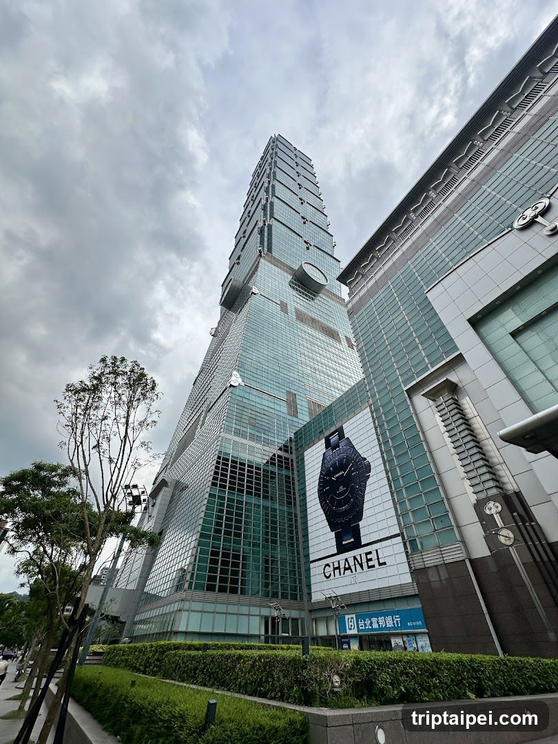 Low-angle view of Taipei 101 skyscraper with glass facade under cloudy sky. Adjacent modern building features large Chanel advertisement on side wall, displaying a watch image above the brand name. Ground level shows green landscaping, streetlights, and partial signage for ‘台北富邦銀行’ (Taipei Fubon Bank) at base of structure.