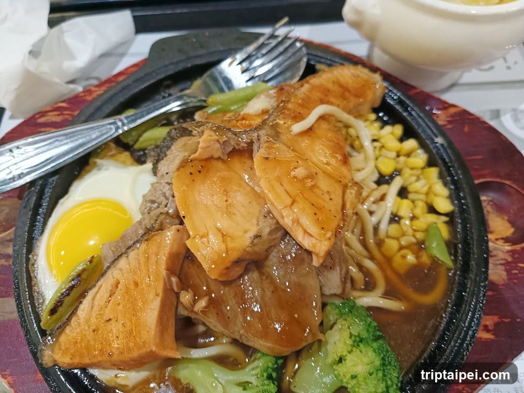 A close-up of a sizzling dish in a black stone pot, featuring sliced chicken breast, corn kernels, bean sprouts, broccoli florets, green onions, and a fried egg with a bright yellow yolk. A fork rests on the edge of the pot. The dish is placed on a red textured placemat.