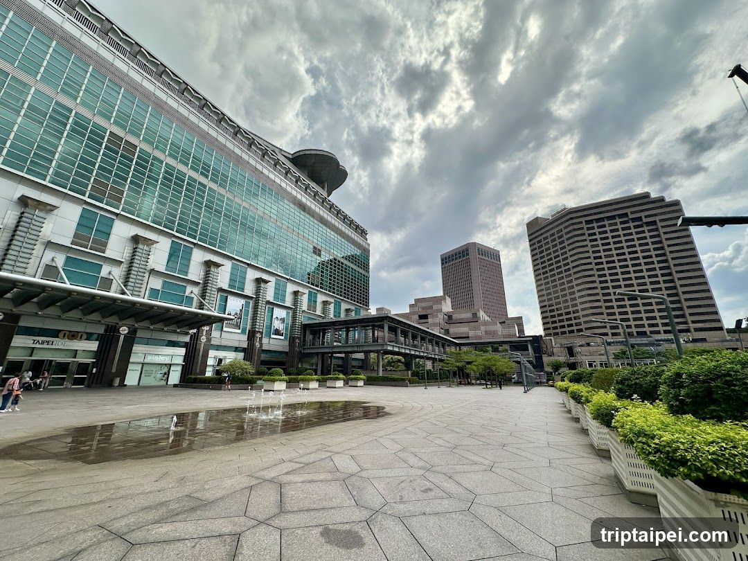 A wide paved plaza in an urban setting, featuring a large glass-fronted building on the left with visible signage including 'TAP' and 'MALL'. A small fountain is active near the foreground. To the right, there are tall office-style buildings under a dramatic, cloud-filled sky. Greenery lines the edges of the walkway. No menus or shop fronts are clearly identifiable.