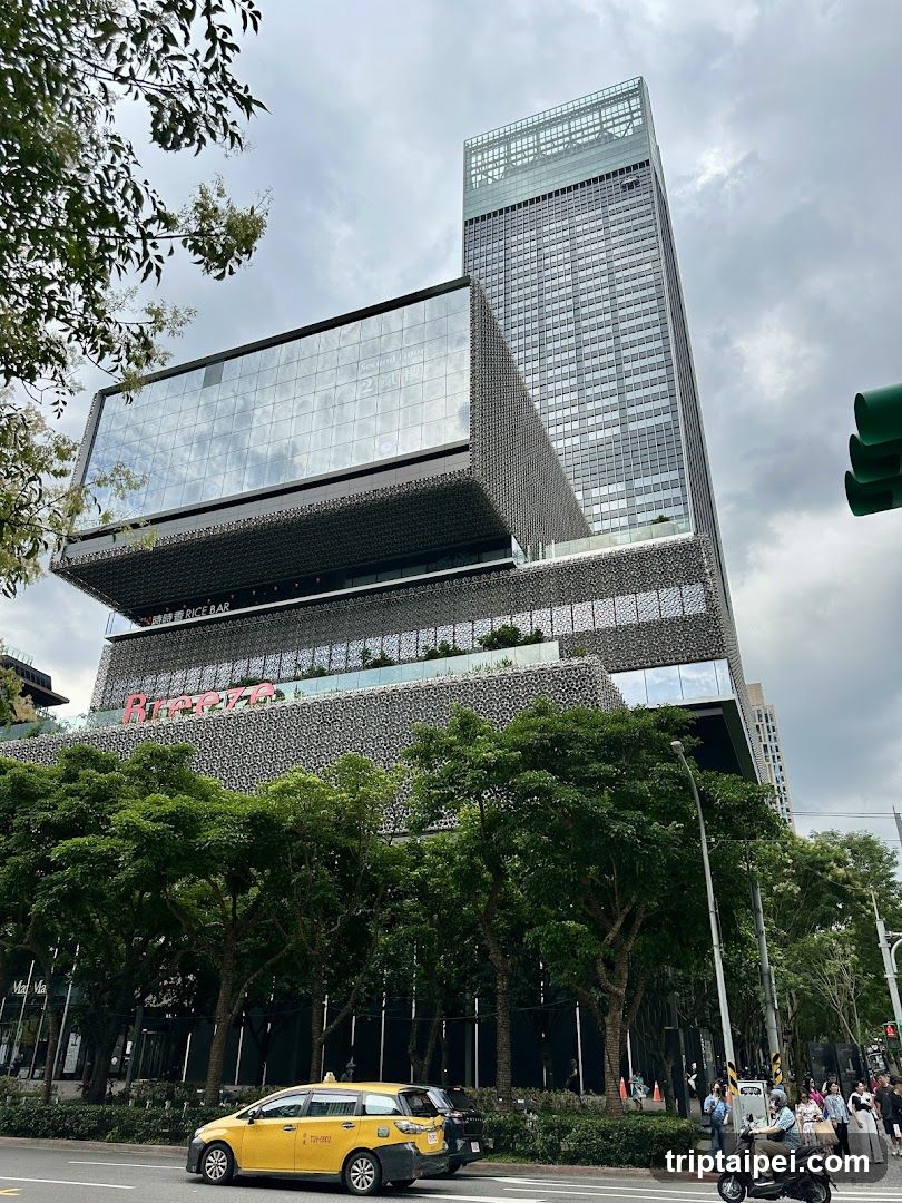 A modern high-rise building with a distinctive architectural design featuring a large glass facade and a textured lower section. The Ritz-Carlton sign is visible on the building's exterior. A yellow taxi and other vehicles are parked in front, surrounded by lush green trees under an overcast sky.
