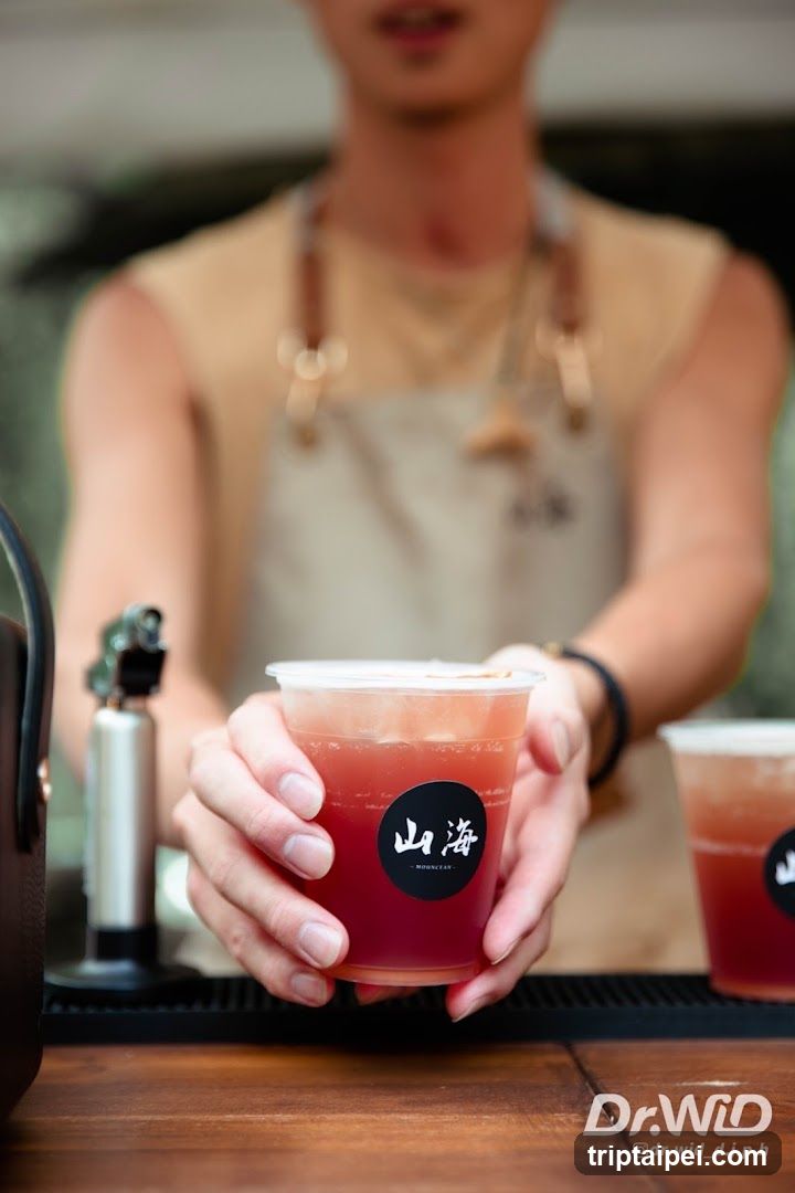A close-up shot of a bartender's hands holding a clear plastic cup filled with a reddish-brown beverage, likely iced tea or juice. The cup has a black circular sticker with white Chinese characters '山海' (Shan Hai). In the background, slightly out of focus, is the upper body of the bartender wearing a beige apron over a sleeveless shirt. To the left, part of a metal shaker and another similar drink are visible on the counter.