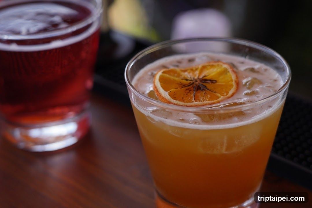 A close-up of two cocktails in glass tumblers, one with an orange slice garnish and the other a dark red drink, both resting on a wooden bar surface.