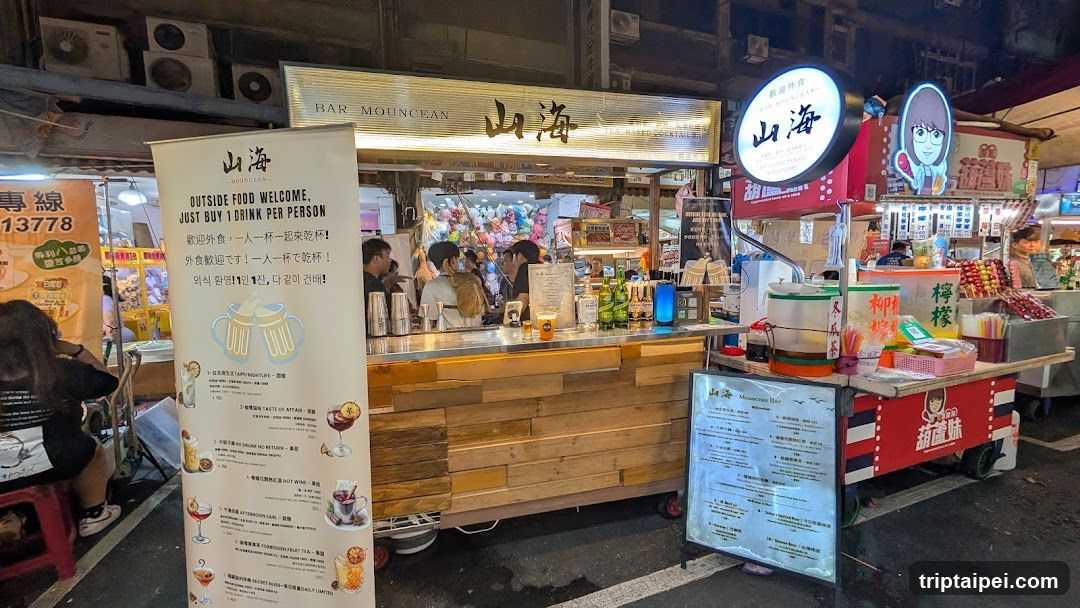 A bustling night market scene featuring a wooden food stall with 'HAR MOUNCEAN' and Chinese characters on its sign. A vertical menu board lists various drinks, while adjacent stalls display colorful snacks and merchandise. The area is lively with customers seated at tables.