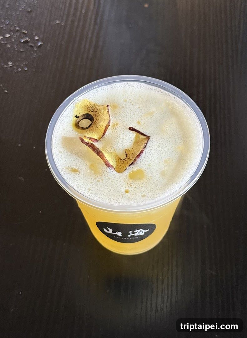 A close-up top-down view of a yellow drink in a clear plastic cup, topped with foam and decorated with two pieces of dried golden-brown fruit (possibly apple or pear). The cup has a black label with white Chinese characters '山海' visible on the side. The background is a dark wooden surface with some water droplets scattered around.
