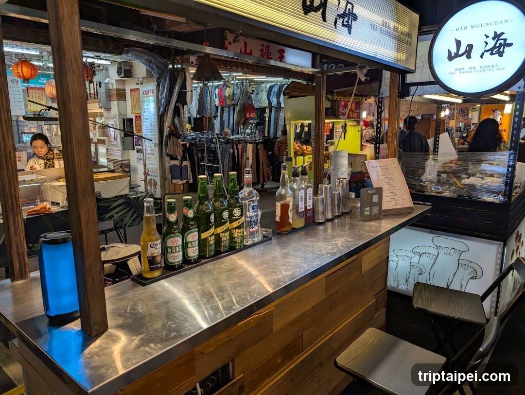 The image shows the interior of a bar or restaurant with a counter displaying various bottles of alcohol, including beer and spirits. A menu board is visible on the right side of the counter. In the background, there are racks of clothing hanging, suggesting a multi-purpose space that might also sell apparel. The lighting is warm, creating a cozy atmosphere.