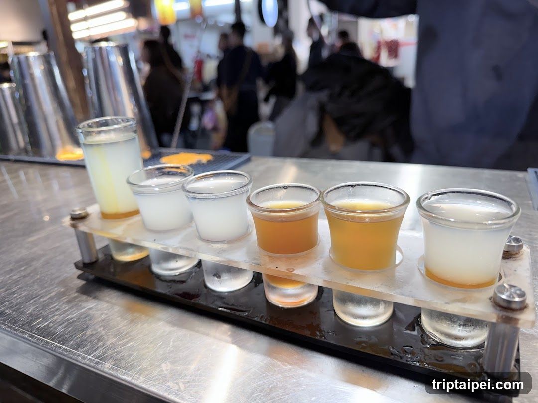 A row of six small shot glasses filled with various liquids, arranged on a metal tray in what appears to be a bar or market setting. The background is blurred but shows people and possibly other stalls.