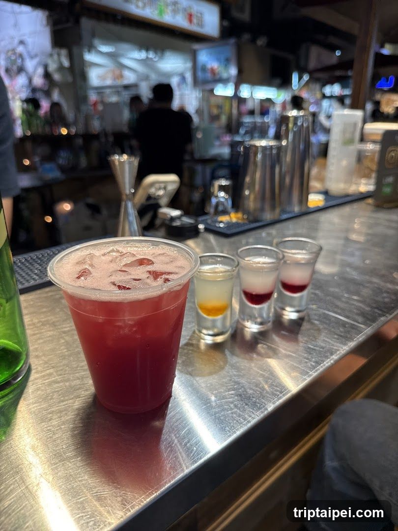 A close-up view of a stainless steel bar counter featuring a large plastic cup filled with pink iced beverage and three small shot glasses containing layered liquids (yellow, red, clear). In the background, blurred figures are visible behind the bar area, along with various bar tools like jiggers and shakers. The setting appears to be an indoor establishment, likely a bar or restaurant.