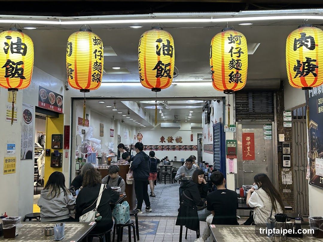 A bustling restaurant entrance featuring bright yellow lanterns with Chinese characters, including '油飯' (rice) and '蚵仔麵線' (oyster noodle). The interior is visible through the open doorway, showing patrons dining at tables.
