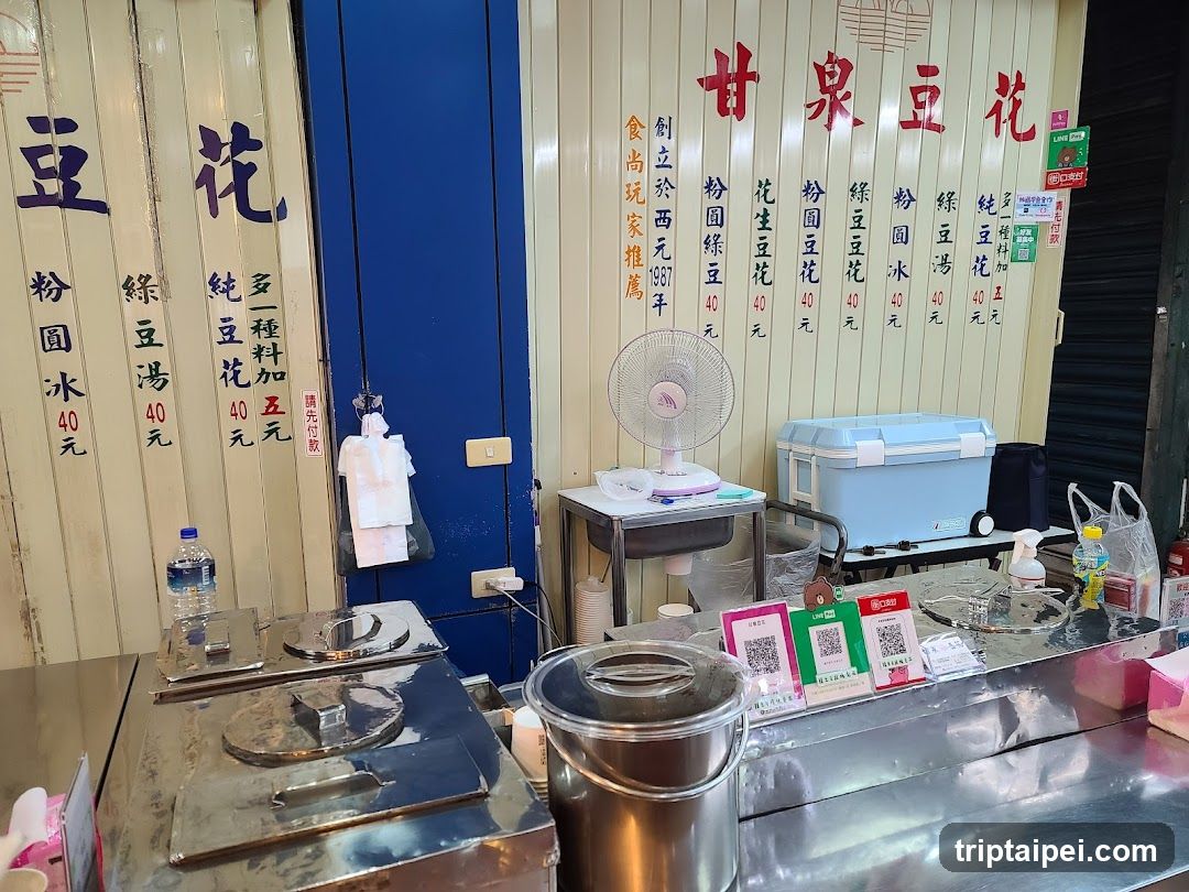 The image shows the exterior of a small food stall or shop with vertical wooden slat walls and blue doors. Prominent red Chinese characters on the wall read '甘泉豆花' (Ganquan Bean Flower). A menu board lists various bean flower products like '粉圓豆花', '綠豆湯', and '純豆花' with prices in NT dollars. There is a metal counter, a fan, a cooler box, and payment QR codes visible.
